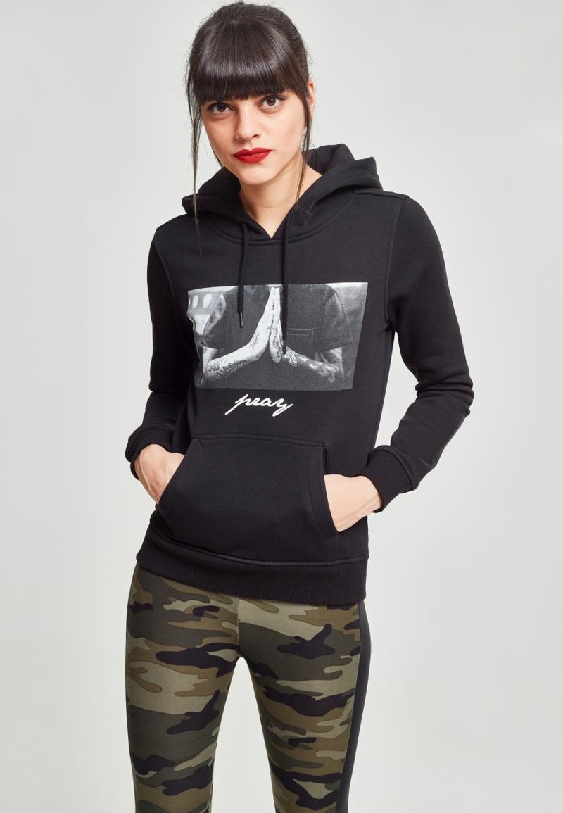 Sudadera negra con capucha y bolsillo frontal, que presenta un gráfico de manos en oración. Combinada con mallas de camuflaje.