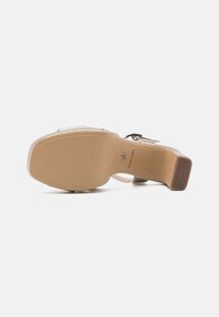 Sandal de tacón beige con un upper liso y brillante, correa de tobillo ajustable y una suela de goma contorneada con patrones de agarre antideslizante.