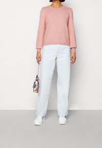 Helles rosa Strickpullover mit langen Ärmeln, kombiniert mit hellblauen Jeans im geraden Schnitt. Ergänzt mit einer mehrfarbigen Handtasche mit Druck und weißen Sneakers.