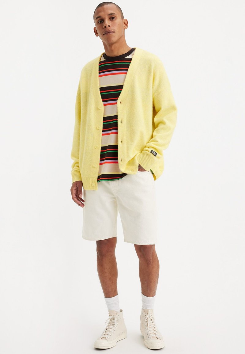Gelbe gestrickte Strickjacke mit V-Ausschnitt, Knopfverschluss, getragen über einem mehrfarbigen gestreiften T-Shirt. Kombiniert mit weißen Shorts und Sneakers.