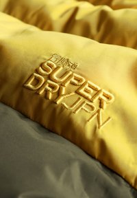 Żółta pikowana tkanina z wyhaftowanym napisem "SUPER DRY JPN" oraz oliwkowozieloną częścią poniżej, podkreślająca wyraźny kontrast kolorów i teksturę.