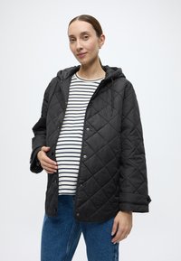 Μαύρο καπιτονέ μπουφάν με κουκούλα, που κλείνει με τρουκ, φορεμένο πάνω από ριγέ πουκάμισο. Το μοντέλο έχει στρογγυλή κοιλιά και στέκεται μπροστά σε απλό φόντο.