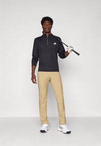 Black zip-up golf pullover, beige golf pants, white shoes with black accents, golfer holding a club, standing against a neutral background.