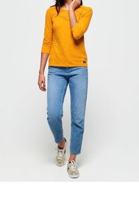 Vrouw in een oranje lange mouwen top, blauwe jeans en beige sneakers die tegen een effen lichte achtergrond loopt.