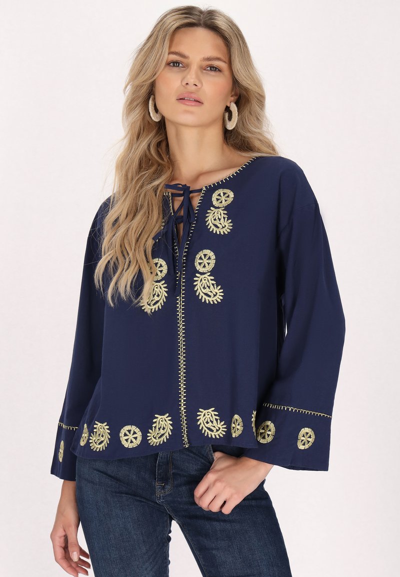 Blusa blu navy con ricami dorati, presenta un collo rotondo, chiusura anteriore con lacci e maniche a campana. Tessuto morbido con dettagli decorativi sull'orlo.