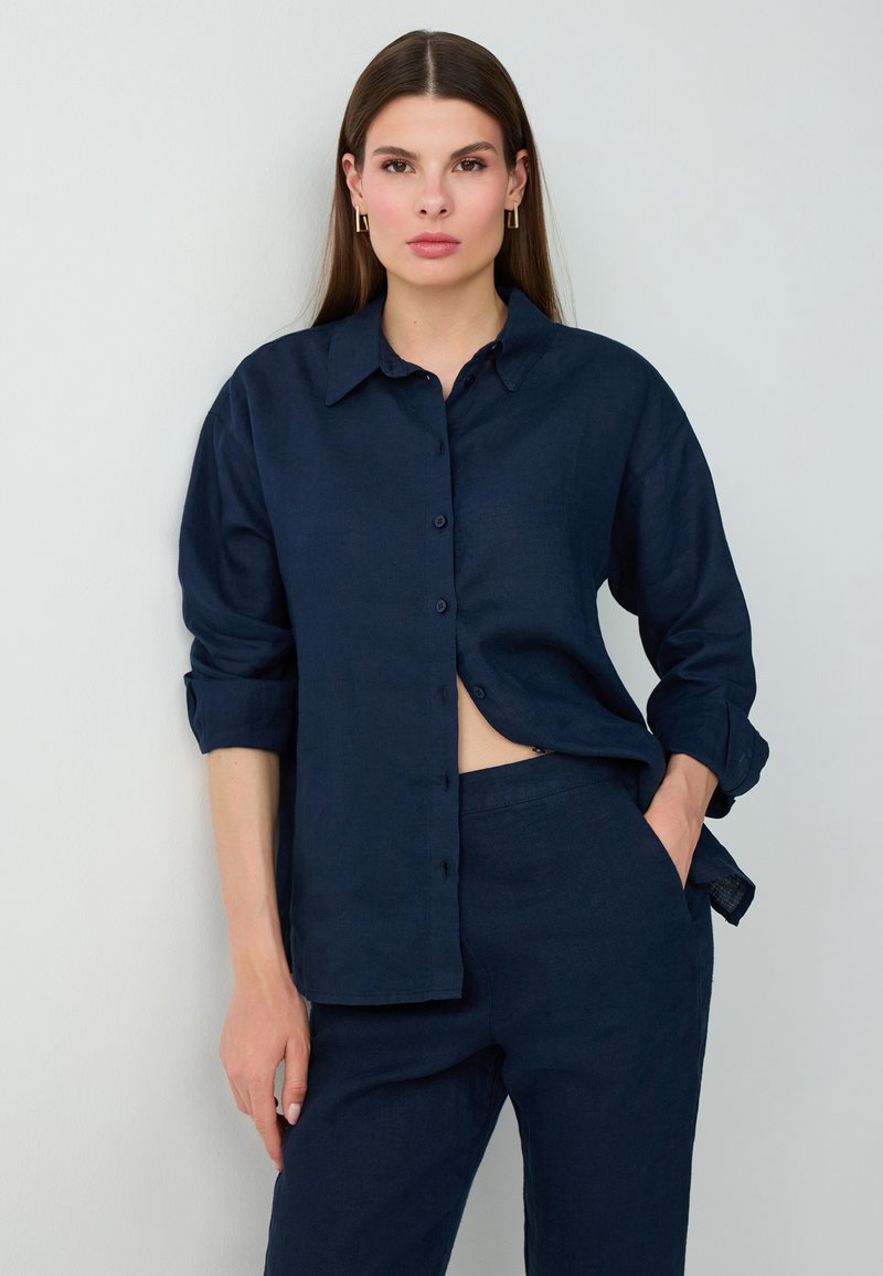 Femme aux longs cheveux bruns porte une chemise bleu marine boutonnée et un pantalon assorti, debout, une main dans la poche, sur un fond clair.