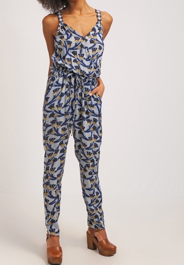 Femme portant une combinaison sans manches à motif floral bleu, jaune et noir, associée à des talons compensés en bois marron.