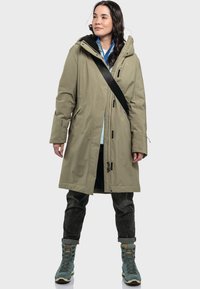 Olivgrüner wasserdichter Parka mit verstellbarer Kapuze, Frontreißverschluss und Schnürverschluss. Kombiniert mit dunklen Jeans und blauen Wanderschuhen.
