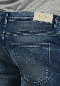 Plave traperice s izblijedjelim završetkom, klasičnim dizajnom s pet džepova i bež kožnom oznakom s natpisom "BLEND" na stražnjoj strani struka.