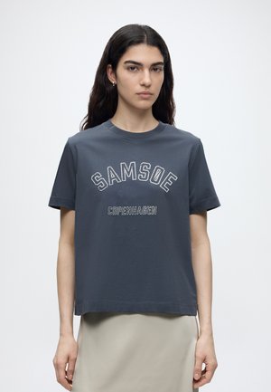 Femme aux longs cheveux foncés portant un t-shirt gris foncé Samsoe Copenhagen et une jupe beige clair, debout sur un fond blanc uni.