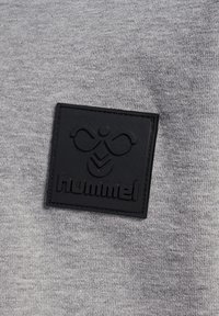 Grå textil med en närbild av en svart gummilogobricka med varumärkesnamnet "hummel" och en stiliserad design i mitten.
