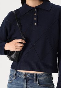 Frau trägt einen marineblauen Strickpullover mit Rautenmuster und vier Knöpfen, hält eine schwarze Leder-Umhängetasche und trägt eine blue Jeans.