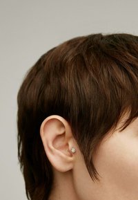 Pendiente de plata con una pequeña piedra preciosa transparente, llevado en el lóbulo, junto a un cabello castaño corto que se mezcle con un fondo claro.