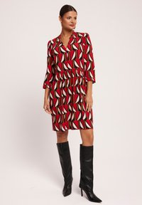 Czerwona sukienka w wzory z długimi rękawami, z geometrycznymi kształtami w kolorze kremowym i czarnym. Noszona z wysokimi na kolana czarnymi botkami na obcasie.