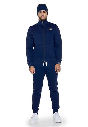 Un homme porte un survêtement bleu marine et un bonnet en maille assorti, avec des baskets blanches, sur un fond blanc uni.