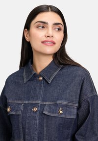 Džínová bunda v tmavomodrej farbe s plochým golierom, vreckami na hrudi a kovovými gombíkmi s kontrastným prešívaním. Hladká textúra a štruktúrovaný dizajn.
