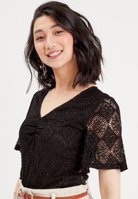 Haut en dentelle noire avec des manches courtes évasées, doté d'un décolleté en V et de motifs floraux complexes. Texture lisse et corsage ajusté avec un détail torsadé.
