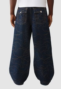 Spodnie dżinsowe z ciemnoniebieską bazą i skomplikowanymi pomarańczowymi abstrakcyjnymi wzorami. Posiadają tylne kieszenie z metalowymi guzikami oraz zwężany krój.