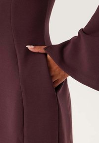 Abito color mogano realizzato in tessuto liscio ed elastico con silhouette aderente e maniche a campana. Tasca laterale visibile con una mano appoggiata all'interno.