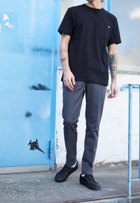 Černé bavlněné tričko s kapsou, šedé elegantní kalhoty, černé tenisky se zlatými detaily, stojící poblíž modrých dveří s grafickým uměním.