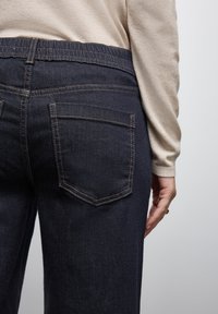 Jeans de denim azul oscuro con una cintura elástica, que cuentan con dos bolsillos traseros y costuras en contraste. La toma en primer plano muestra la textura y los detalles del diseño.