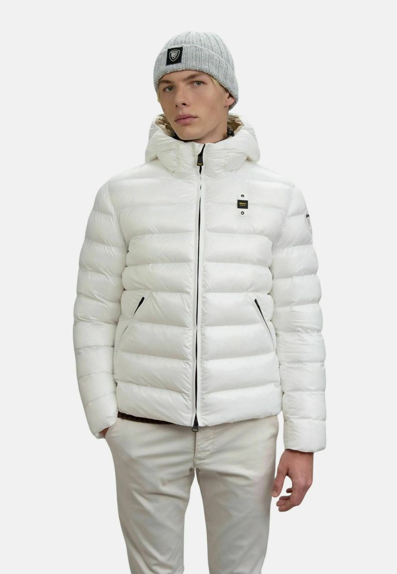 Un giovane uomo indossa una giacca puffer bianca con cappuccio, pantaloni beige chiaro e un berretto di maglia grigio con una toppa nera.