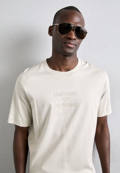 Μπεζ κοντομάνικο μπλουζάκι με ανάγλυφο λογότυπο "EMPORIO ARMANI". Το μοντέλο φορά σκούρα γυαλιά ηλίου με γυαλιστερό χρυσό σκελετό.