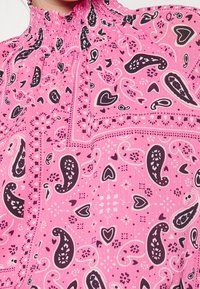 Blouse rose avec un col smocké, ornée d'un motif cachemire en violet foncé et blanc. Tissu texturé avec une drapé fluide et un détail complexe.