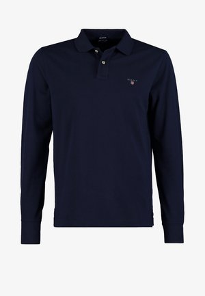 Marineblaue Langarm-Poloshirt, schwarze Jeans und weinrote Sneakers. Die Person trägt eine schwarze Umhängetasche mit braunen Akzenten.