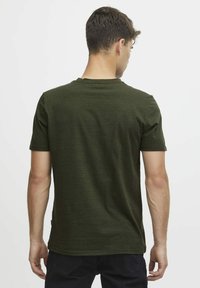 Mørkegrønn t-skjorte med korte ermer, rund hals, strukturert stoff og rett linning, båret av en person som vender ryggen til betrakteren.