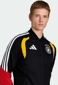 Veste de survêtement noire avec des accents jaunes et rouges, comportant des bandes blanches sur les épaules et un logo brodé avec cinq étoiles.