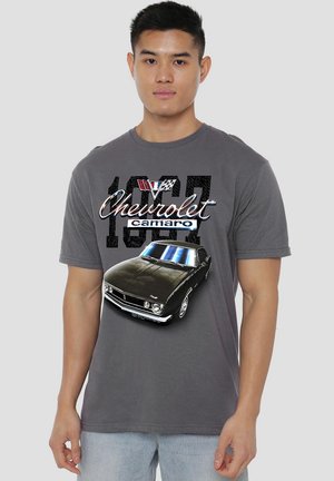 Junger Mann, der ein graues T-Shirt trägt, das ein schwarzes Grafiken eines Chevrolet Camaro von 1969 mit Text und Rennflaggen zeigt.