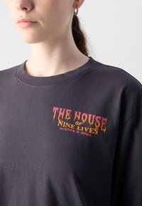 Mørk grå bomulls-T-skjorte med rund hals og korte ermer, med et fargerikt grafisk trykk: "THE HOUSE of NINE LIVES" i stiliserte bokstaver.