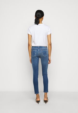 Jean skinny bleu avec poches arrière, associé à un t-shirt blanc uni. Les jeans ont un délavage moyen et une texture lisse.