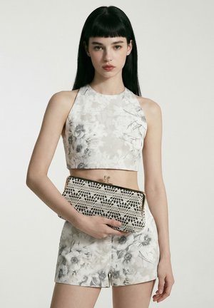 Jeune femme aux longs cheveux noirs porte un crop top sans manches à motif floral et un short, tenant une pochette noire et blanche à motifs.