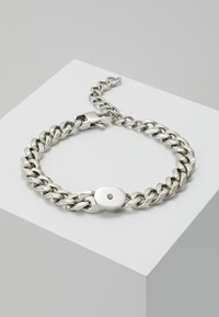 Helohopeinen ketjukaulakoru, jossa on soikea lukko ja pieni keskuskivi. Sisältää toisiinsa linkitettyjä renkaita, joilla on sileä ja kiiltävä pinta.