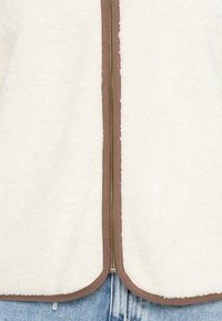Witte fleece vest met een bronzen rits en een bruis bruine rand langs de randen, met een zachte textuur en een afgeronde vorm aan de onderkant.
