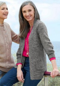 Zwei lächelnde Frauen im Freien am Meer, eine in einem rosa Hemd mit grauem Cardigan, die andere in einem beigen Kabelstrickpullover.