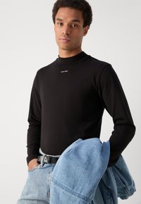 Must mustariga T-särk kõrge kaelusega, millel on väike valge "Calvin Klein" logo. Kombineeritud helesiniste teksadega.