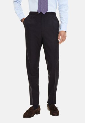 Mann trägt dunkle, maßgeschneiderte Hose, hellblaues Hemd, gemusterte Krawatte und braune Wildleder-Loafer mit Quasten vor einfachem Hintergrund.