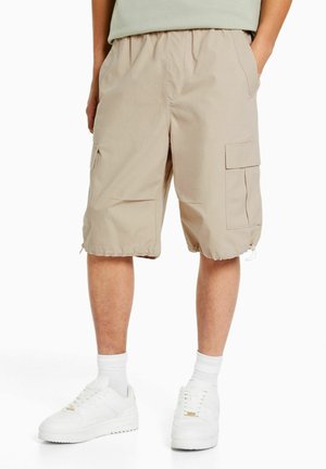 Shorts cargo beige clair avec taille élastique, poches latérales et détails en zip à l'ourlet, fabriqués dans un tissu lisse et léger.