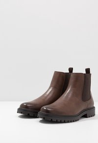 Botas Chelsea de cuero marrón con paneles laterales elásticos, un acabado texturizado y una suela de goma negra rugosa para mayor tracción.