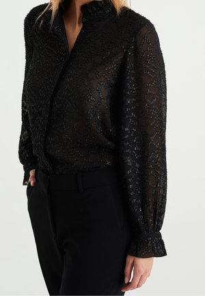 Vrouw draagt een doorschijnende zwarte blouse met gestructureerde patronen en opgeblazen mouwen, gecombineerd met een zwarte broek, hand in zak, op een effen achtergrond.