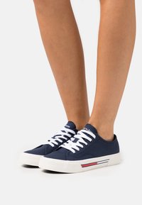 Navyblå canvas sneakers med vita snören, en gummisula och en röd- och vitrandig accent på sidan. Rundad tådesign.