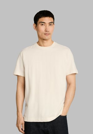 Jonge man met kort zwart haar, gekleed in een effen gebroken wit T-shirt met korte mouwen en donkere broek, staand voor een grijze achtergrond.