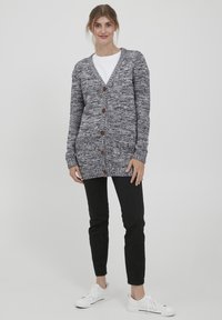 Cardigan long en maille épaisse gris et noir avec cinq boutons marron, décolleté en V, et deux poches avant, associé à un pantalon noir et des baskets blanches.