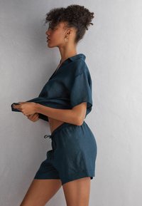 Produkter: Tvådelad outfit i mörk teal, tillverkad av linne. Innehåller en lös kortärmad skjorta med krage och shorts med dragsnöre. Slät textur.