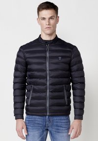 Chaqueta acolchada negra con acolchado horizontal, cierre frontal y cuello alto. Cuenta con dos bolsillos laterales con cremalleras y logo en el pecho.