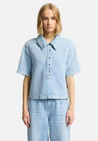 Lys blå denimskjorte med stor krave, korte �ærmer, fem-knaps lukning og cropped søm. Blød tekstur med diskret falmning.