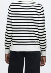 Personne portant une chemise blanche à manches longues avec des rayures horizontales noires et un pantalon noir, vue de dos.